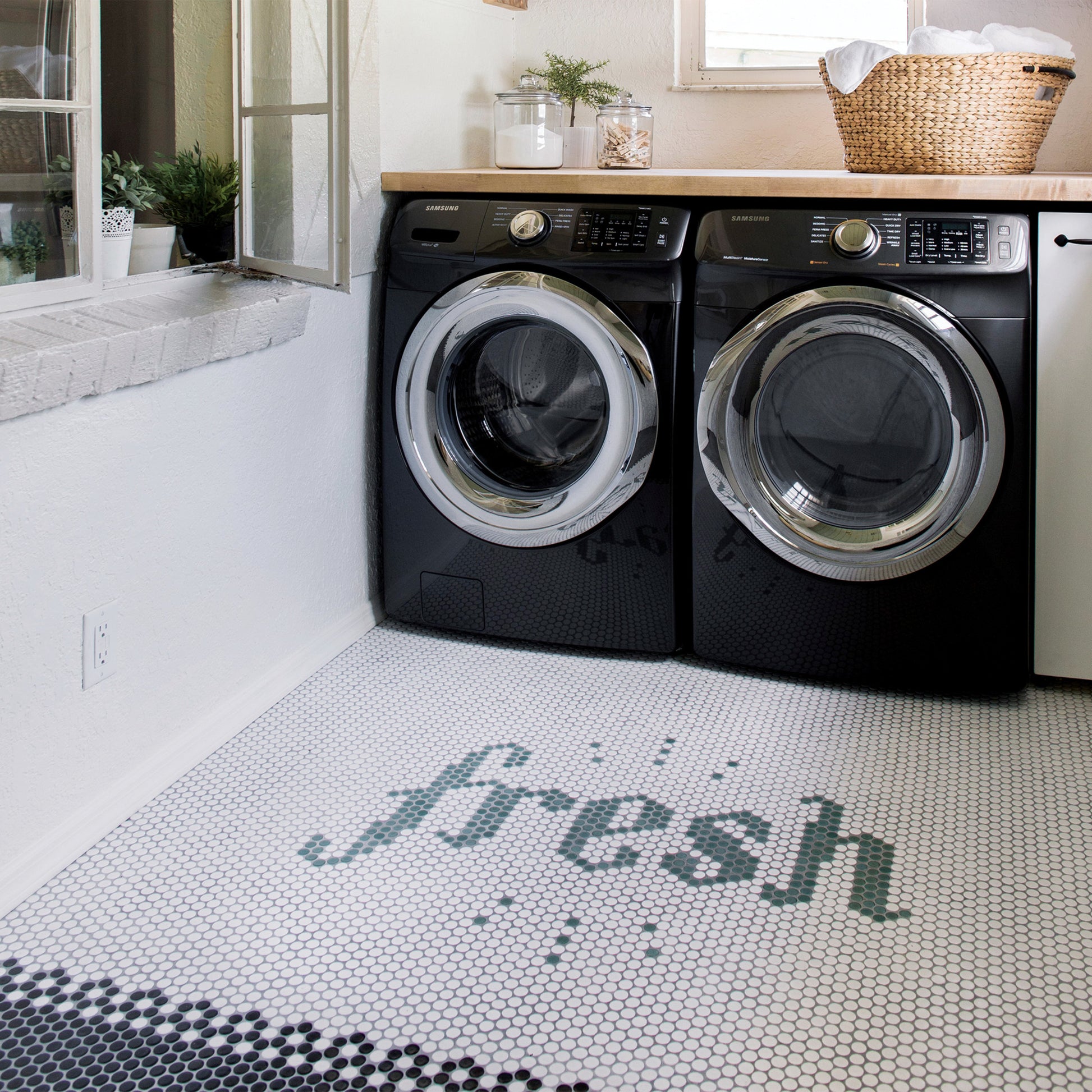 A laundry room with black and silver washer and dryer. The floor is all white penny tile leading into black penny tile. The middle of the floor, in Shale green penny tile, spells out Fresh