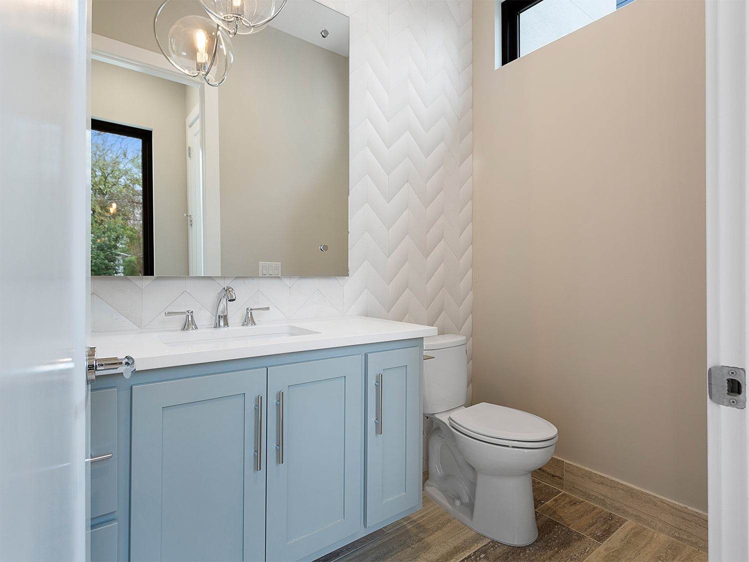 Modern bathroom, with light blue cabinets to the left with a white marble countertop, with silver hardware and a full wall of 10.5" x 11" white natural stone tile in a chevron pattern. 