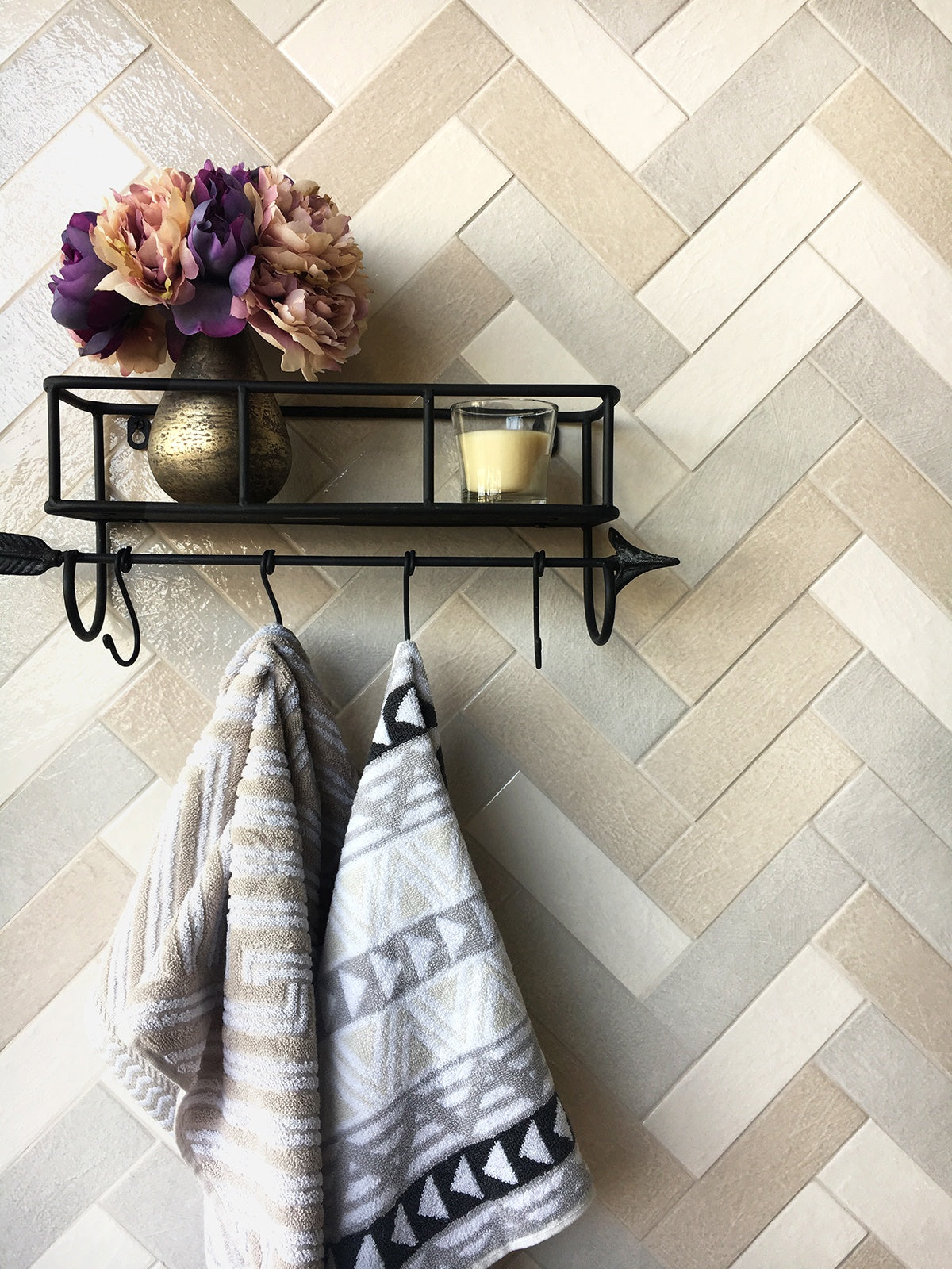 A black, metal towel rack in the shape of an arrow with a shelf placed on top with purple and pink flowers in a vase, next to a clear glass candle. The shelf is affixed to 2.5" x 8" Abitare La Terra wall tile. The tile layout is in the herringbone style, with multiple colored tile. The colors include antracite, bianco, and sabbia.