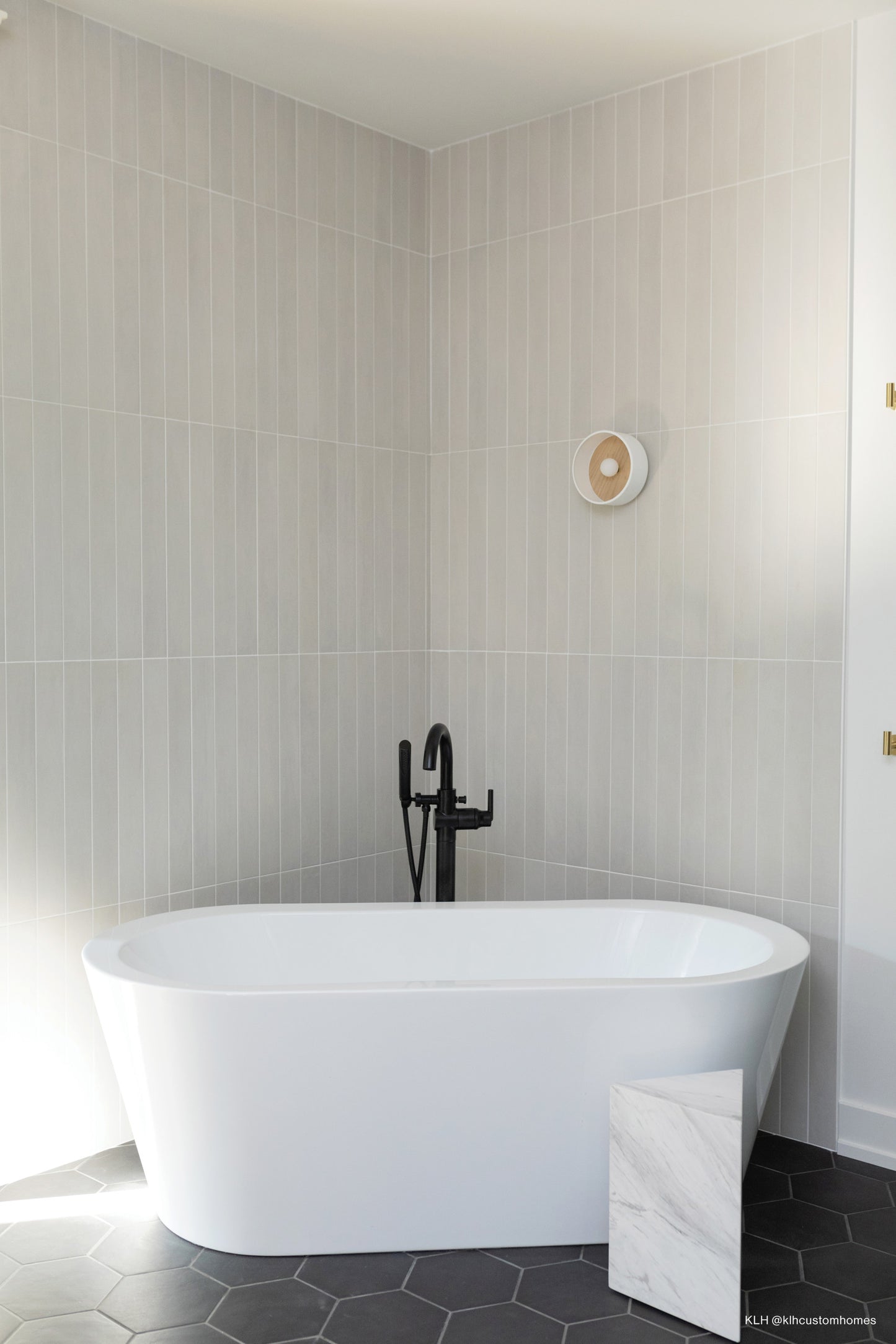 White matte field tile laid vertical on a wall behind a white bathtub. Flooring is the black hexagon field tile from Allora.